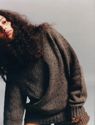 Portrait of a young woman with long brown curly hair, wearing a brown knit sweater and brown striped skirt against a grey background.