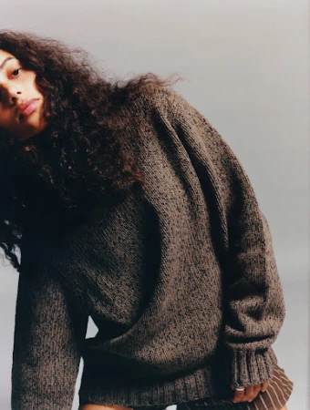 Portrait of a young woman with long brown curly hair, wearing a brown knit sweater and brown striped skirt against a grey background.