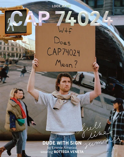 Man holding sign in a public space with buildings and people in background, street crossing signal visible, fashionable street style.