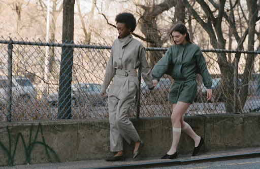 Two women in fashionable jumpsuits walking hand-in-hand on a city sidewalk with trees and buildings in the background on a sunny day.
