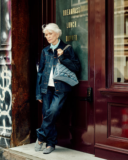 Mature woman in denim jacket and jeans standing in front of a restaurant door in an urban environment, carrying a quilted denim bag.