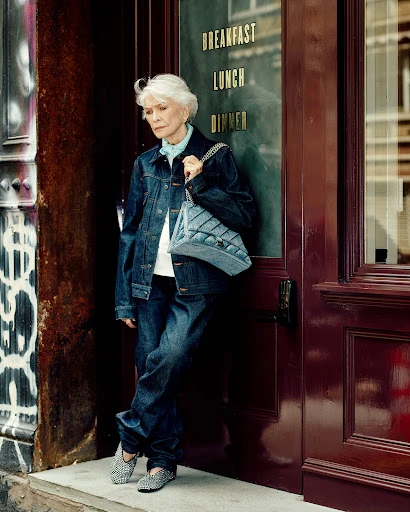 Mature woman in denim jacket and jeans standing in front of a restaurant door in an urban environment, carrying a quilted denim bag.