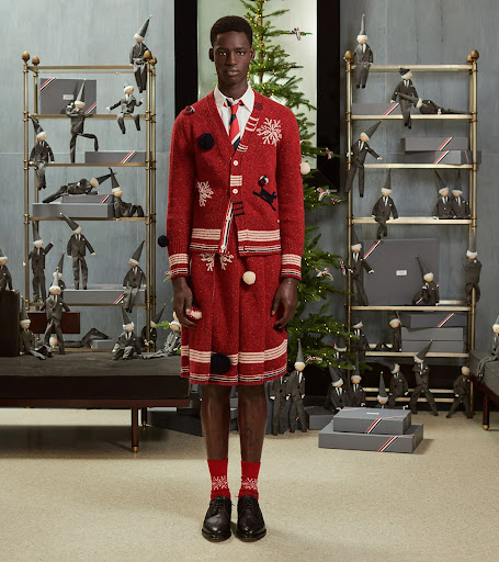 Editorial fashion shot of a Black male model in a red knit cardigan and skirt, accessorized with a tie and socks, posed indoors.