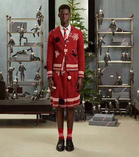 Editorial fashion shot of a Black male model in a red knit cardigan and skirt, accessorized with a tie and socks, posed indoors.