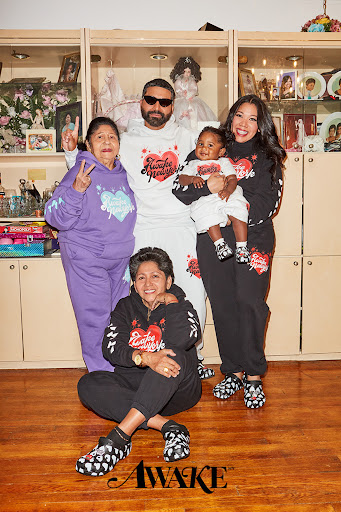 Group portrait of five people wearing matching sweatsuits with heart graphics in a home setting, with a display cabinet in the background.