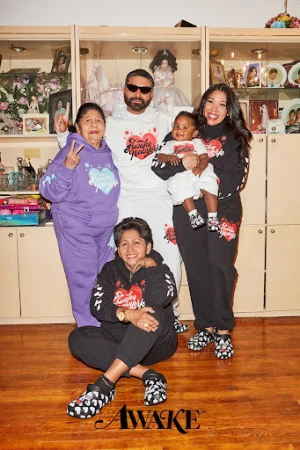 Group portrait of five people wearing matching sweatsuits with heart graphics in a home setting, with a display cabinet in the background.