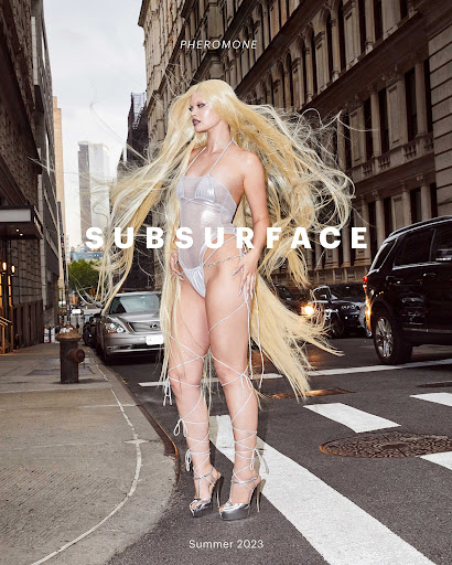 Fashion shoot on city street featuring woman with long blonde hair wearing light silver bodysuit, and silver high heels on crosswalk.