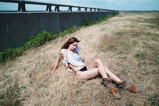 Woman in white top and boots seated in dry grass outdoors by concrete wall