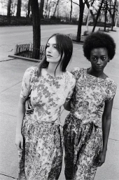 Two female models in patterned outfits walking together on an urban sidewalk lined with bare trees.
