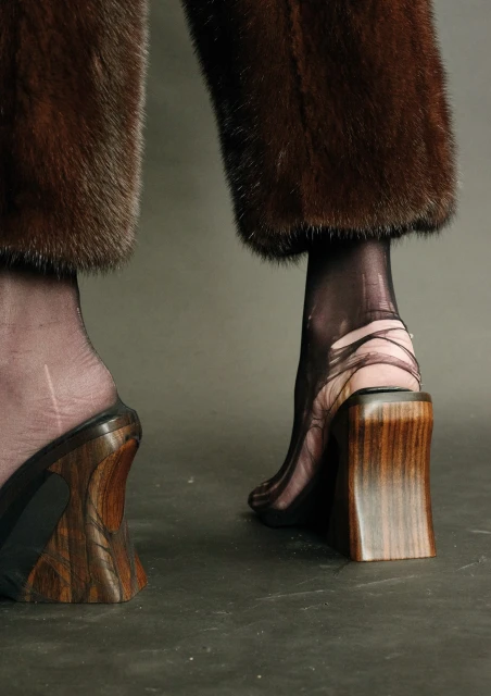 Close-up of woman's legs with brown fur pants, black sheer tights, and wood block heel sandals, with neutral gray background.