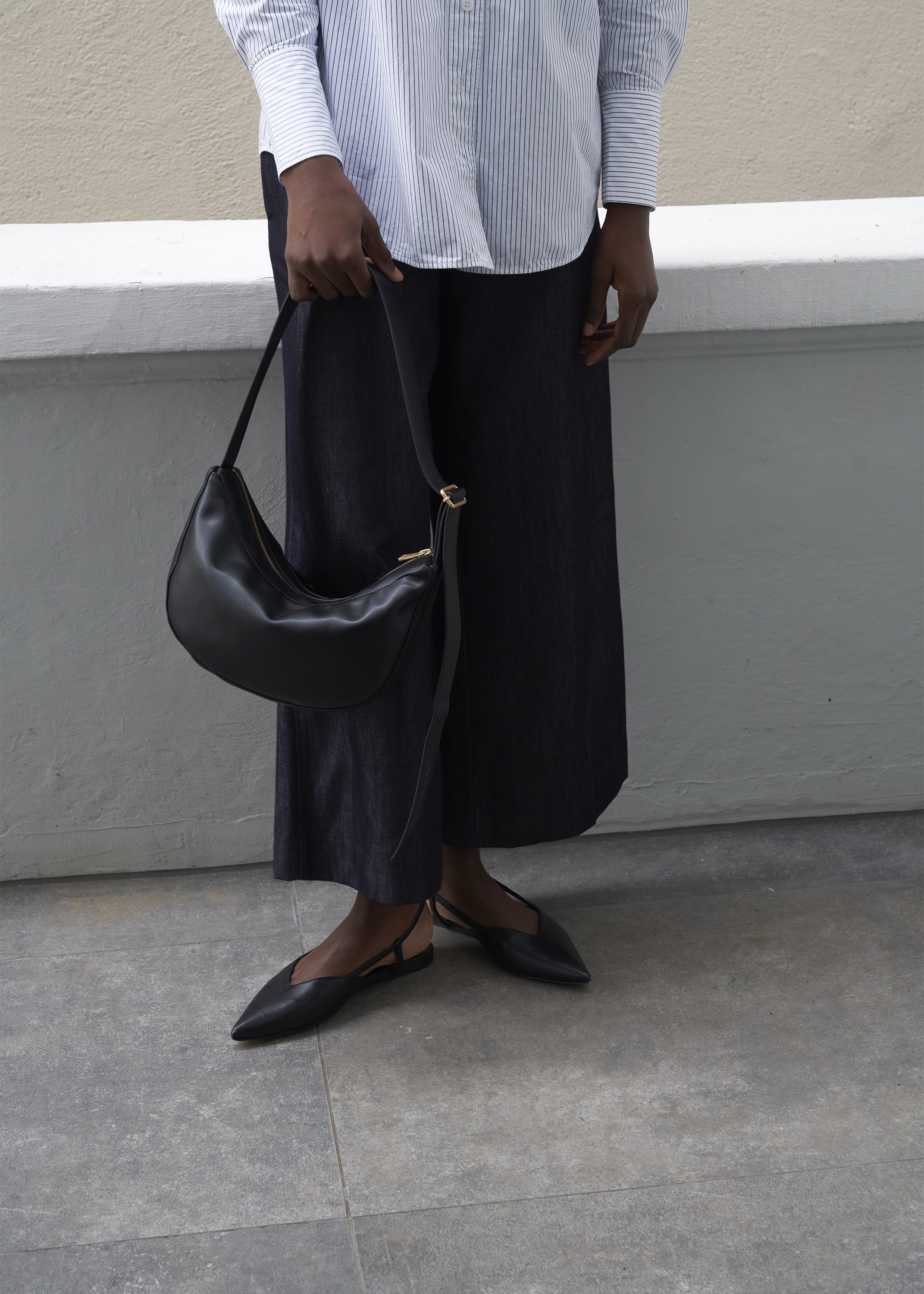 Fashionable outfit featuring a woman holding a black leather purse, paired with a striped shirt, dark pants, and pointed black shoes, outdoors.