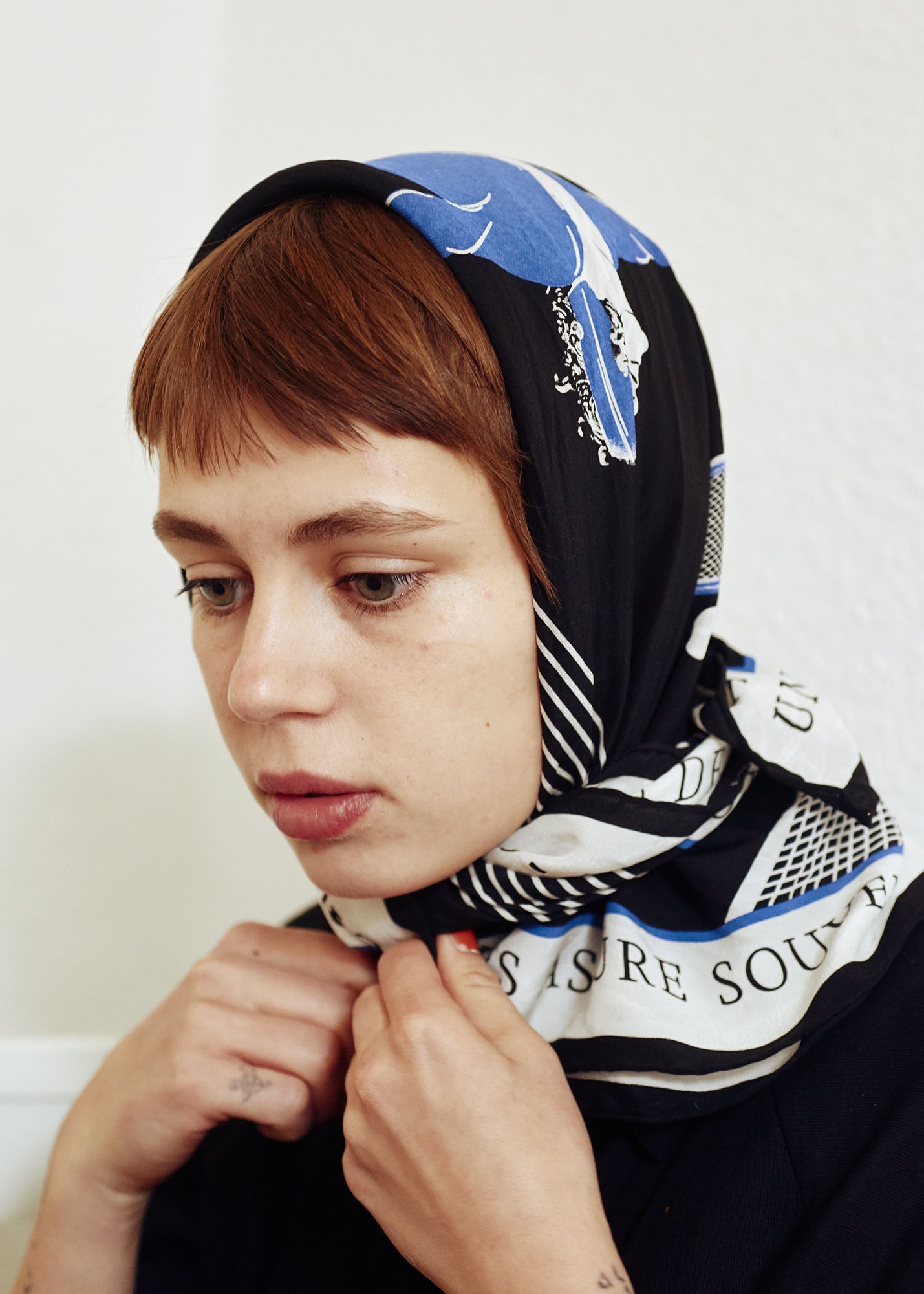 Close-up portrait of a young woman with red-brown hair wearing a black, white, and blue printed headscarf against a white background.