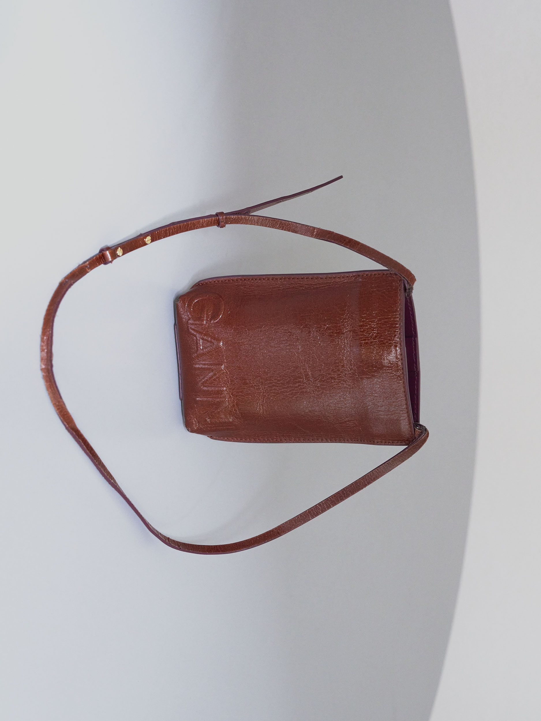 Brown leather Ganni crossbody bag with thin strap and logo detail against a minimalist grey and white background.