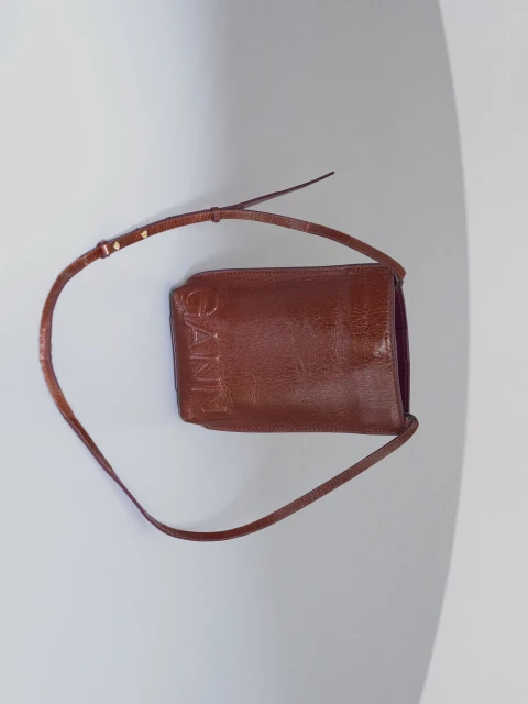 Brown leather Ganni crossbody bag with thin strap and logo detail against a minimalist grey and white background.