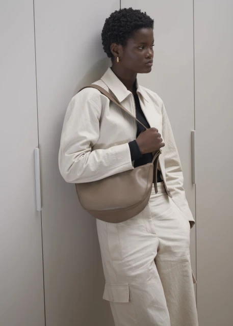 Fashion shot of an African-American woman wearing a beige jacket and cargo pants with a handbag, standing against a grey wall.
