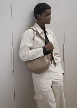 Fashion shot of an African-American woman wearing a beige jacket and cargo pants with a handbag, standing against a grey wall.
