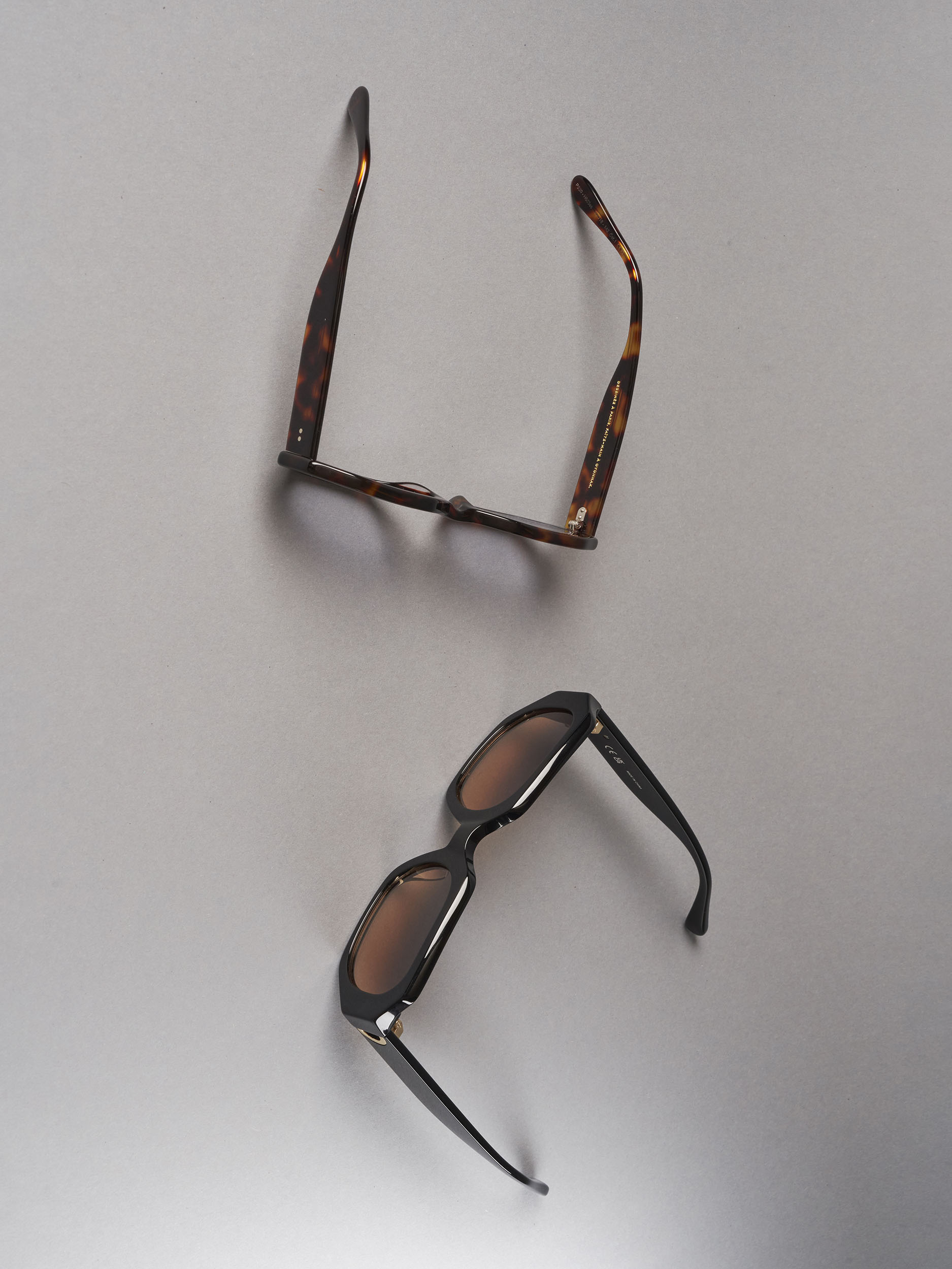 Two pairs of fashion sunglasses are placed on a light grey surface, one with brown frames and the other with black frames.