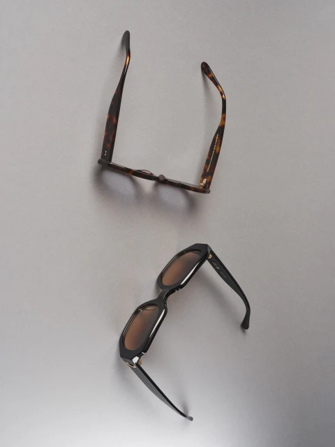 Two pairs of fashion sunglasses are placed on a light grey surface, one with brown frames and the other with black frames.
