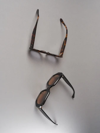 Two pairs of fashion sunglasses are placed on a light grey surface, one with brown frames and the other with black frames.