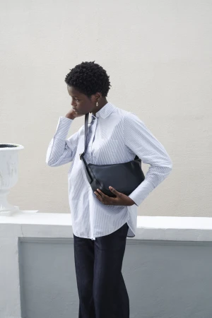 Stylish woman wearing a blue striped shirt and dark jeans, carrying a black leather shoulder bag against a neutral background.