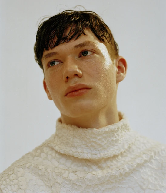 Headshot of a light-skinned adult male with short reddish-brown hair wearing a textured white mock turtleneck, looking left against a white backdrop.