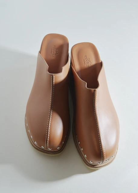 Pair of brown leather clogs with white stitching and silver stud accents displayed on a white surface, studio product shot, fashion accessory.