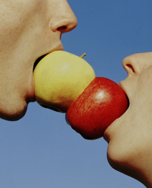 Close-up of two people biting into a yellow and red apple, set against a clear blue sky. Focus on the fruit and the individuals' mouths.