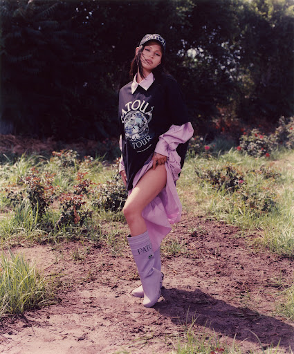 A young woman poses outdoors wearing a black graphic sweatshirt, lilac boots and cap, with a light pink skirt peeking out from beneath the sweatshirt.