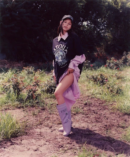 A young woman poses outdoors wearing a black graphic sweatshirt, lilac boots and cap, with a light pink skirt peeking out from beneath the sweatshirt.