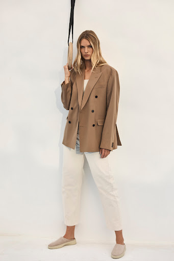 A woman poses in an oversized tan blazer and white pants, standing indoors with a neutral background, holding an exercise ring overhead.