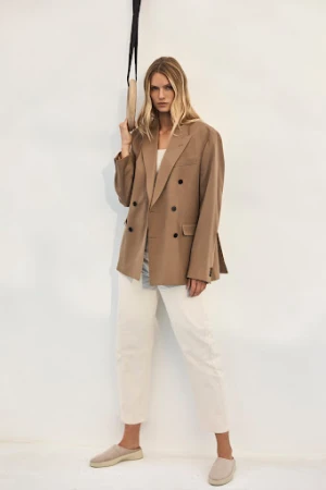 A woman poses in an oversized tan blazer and white pants, standing indoors with a neutral background, holding an exercise ring overhead.