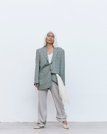 Elderly woman with long white hair in an oversized grey plaid blazer and matching pants poses in front of a white wall.