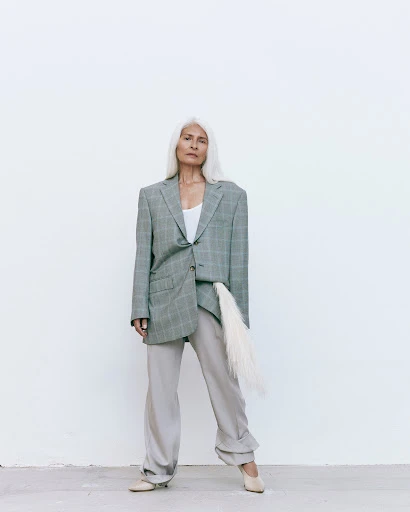Elderly woman with long white hair in an oversized grey plaid blazer and matching pants poses in front of a white wall.