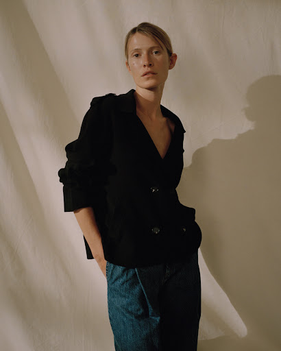 Model in black jacket and blue denim jeans standing against draped fabric backdrop