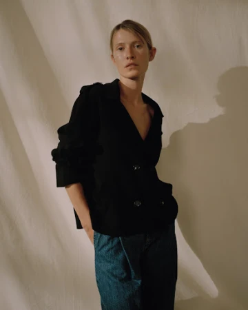 Model in black jacket and blue denim jeans standing against draped fabric backdrop