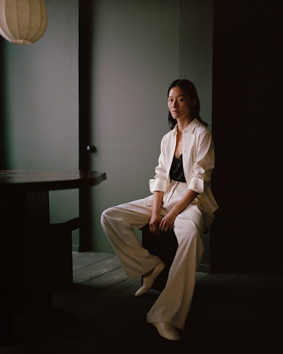 Stylish Asian woman in cream suit sits indoors, shot in moody, low lighting, with focus on fashion and elegant, neutral colors.