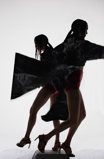 Two women in motion, wearing red shorts, black high heels, and black tops with a white background showcasing fashion and dynamic movement.