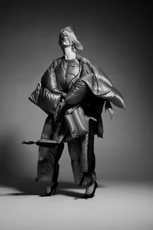 Model with hair blowing across face wearing an oversized quilted puffer coat against a grey gradient backdrop