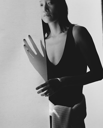 Monochrome portrait of an Asian woman in a black tank top, subtly posed behind a white board, showcasing hands and fabric texture.