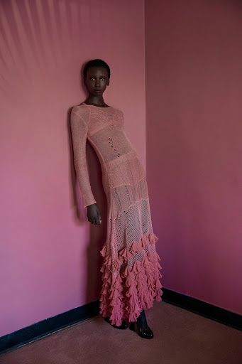 A black female model wearing a long pink crocheted dress posing in a pink room with a shadow.