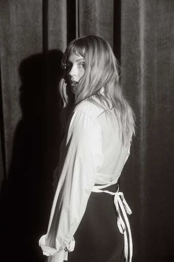 Black and white portrait of a woman with long hair in a white shirt with curtains in the background, looking back over her shoulder.