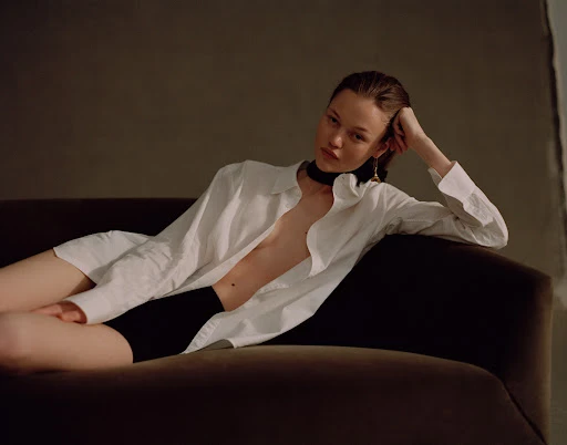 Model lying on a dark sofa wearing an unbuttoned white shirt and black shorts with hair styled by Kelly Peach.