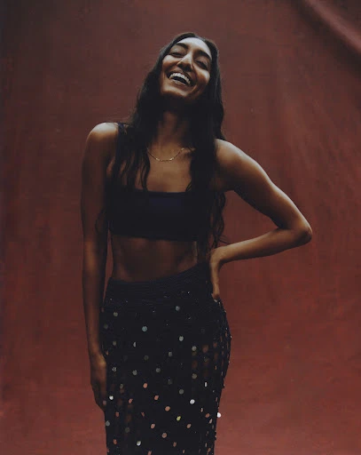 Smiling woman with long dark hair in a dark two-piece outfit against a solid red backdrop, fashion pose, and a happy expression.