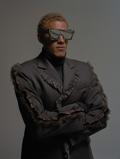 Studio shot of a fashionable adult male with blond hair wearing sunglasses and a black fringe suit, posing with arms crossed against a grey backdrop.