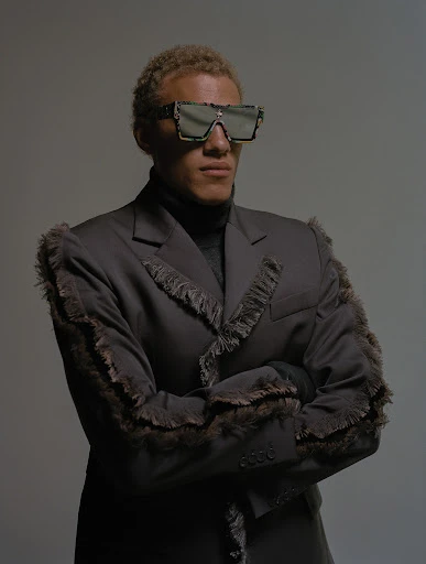 Studio shot of a fashionable adult male with blond hair wearing sunglasses and a black fringe suit, posing with arms crossed against a grey backdrop.