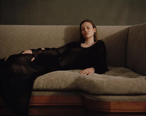 An adult woman lies on a light gray couch wearing a black, long-sleeved sheer top with a dark gray wall in the background.