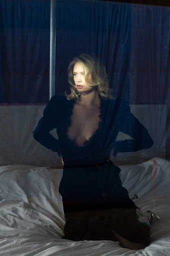 A woman with blonde hair, wearing a black dress, poses while sitting on a white bed in front of a window and blue curtains.