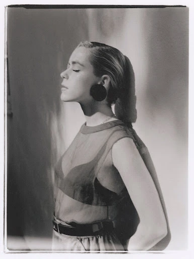 Monochrome portrait of a blonde woman with eyes closed, wearing a sheer top, facing left with a soft light, in a photo studio.