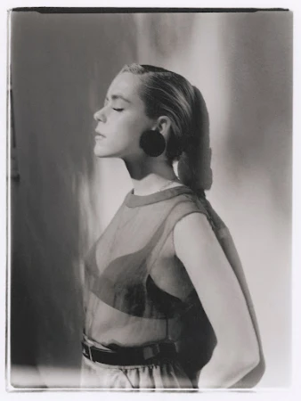 Monochrome portrait of a blonde woman with eyes closed, wearing a sheer top, facing left with a soft light, in a photo studio.