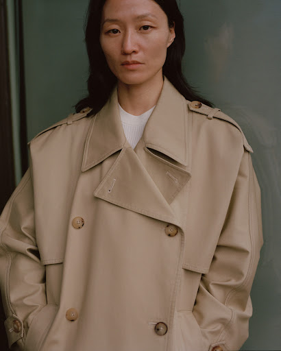Portrait of a woman in a tan trench coat against a neutral green background, looking directly at the camera with a serene expression.